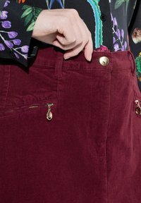 Boutonnage du haut des pantalons en velours côtelé bordeaux taille haute avec des tirettes dorées, et une chemise noire à fleurs rentrée.