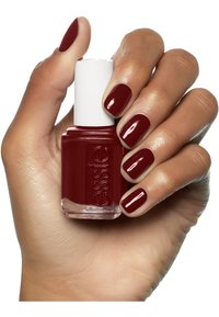 Smalto per unghie rosso scuro in una bottiglia di vetro trasparente, con un tappo bianco. Mano che mostra unghie curate, anch'esse dipinte di rosso scuro. Finitura liscia e lucida.