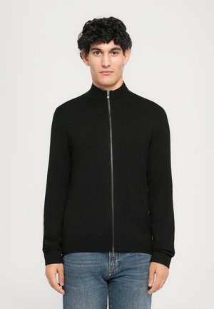 Jovem com cabelo escuro e encaracolado, a usar um casaco preto de fecho e jeans azuis, de frente contra um fundo claro.