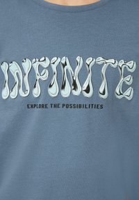 Katoenen t-shirt in gedempt blauw, met glanzende, golvende zilveren tekst "INFINITE" en zwarte tekst "EXPLORE THE POSSIBILITIES" eronder.