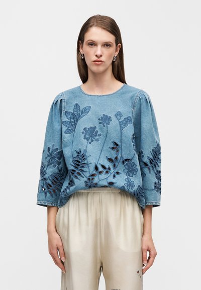 Blouse en denim avec une broderie florale complexe bleu foncé et des détails ajourés. Dotée d'un col rond et de manches trois-quarts.
