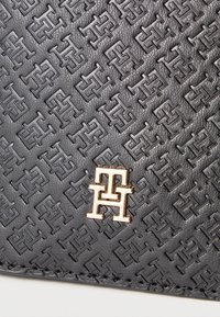 Μαύρο δερμάτινο πορτοφόλι με ανάγλυφο σχέδιο από διασυνδεδεμένα λογότυπα και χρυσή μεταλλική διακοσμητική λεπτομέρεια στο κέντρο. Η υφή είναι λεία με διακριτική λάμψη.