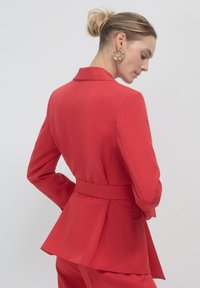 Blazer rosso su misura con design a peplo, colletto singolo e cintura in vita. Tessuto liscio, tasche posteriori e spalle strutturate.