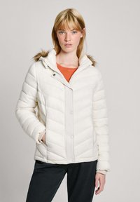 Witte pufferjas met chevronstiksel, capuchon met faux bontafwerking en drukknoopsluiting. Draagt over een oranje shirt, gecombineerd met donkere broek.
