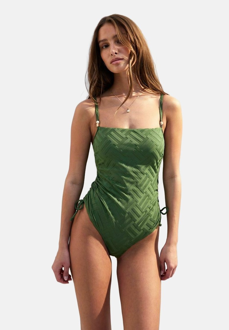 Maillot de bain une pièce vert avec un motif en croix texturé, des bretelles réglables et des liens sur les côtés. Tissu lisse avec une silhouette ajustée.