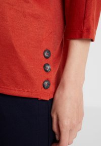 Rotes Strickoberteil mit drei dunklen Knöpfen am Saum. Glatte Textur mit kurzen Ärmeln, tailliertem Design und leicht abgerundetem Saum.