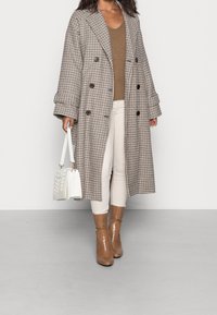 Double-breasted geruite jas in beige en bruin, gecombineerd met een bruine trui, witte broek en beige lakleren enkellaarzen. Witte handtas met geweven ontwerp.