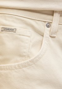 Κοντινό πλάνο σε τσέπη μπεζ τζιν με μεταλλικό εξάρτημα και μικρή υφασμάτινη ετικέτα με τη λέξη "LINDBERGH" στην ραμμένη άκρη.