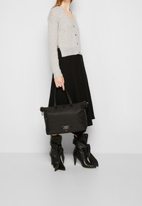 Schwarze Handtasche mit strukturiertem Muster und zwei Griffen. Graue Strickjacke mit Knöpfen und V-Ausschnitt, kombiniert mit einem schwarzen plissierten Rock und knielangen Stiefeln.