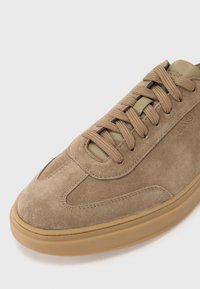 Suede beige sneaker with rubber sole, lace-up design, padded tongue, and small logo detail. Rounded toe and textured surface.