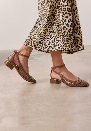 Sandalias de tacón trenzadas en marrón con una correa al tobillo y tacón ancho. Presentan una punta afilada y sutiles acentos dorados. Falda con estampado en el fondo.