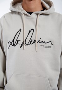 Sudadera color gris claro hecha de una suave mezcla de algodón, con un gran logo negro bordado en el pecho y un bolsillo tipo canguro en la parte delantera.