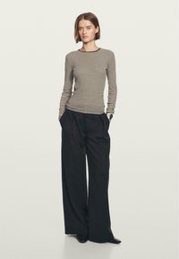 Donna con capelli corti castani, che indossa una maglietta a maniche lunghe beige e nera a righe, pantaloni neri larghi e scarpe nere, in piedi con le mani nelle tasche.