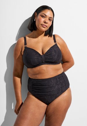 Žena s tmavými vlasmi po plecia, oblečená v čierne bikiny s vysokým pásom a kvetinovým vzorom, stojaca pri bielej stene so tieňom.