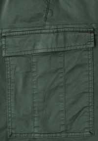Poche en tissu vert avec un rabat rectangulaire fixé par des coutures, montrant des coutures détaillées et de légères rides dans le matériau.