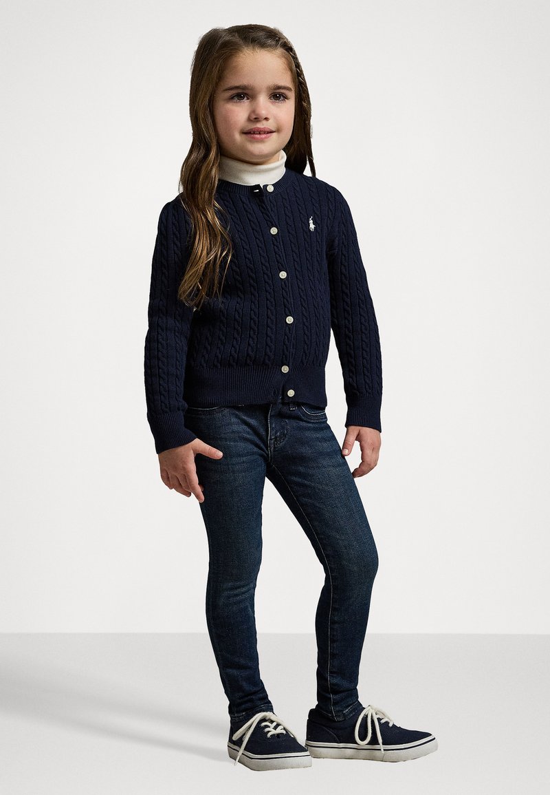 Marineblaue Kabelstrick-Cardigan mit Rundhalsausschnitt, Knöpfen und kleinem Logo, kombiniert mit dunkelblauen Jeans und marineblauen Sneakers mit weißen Sohlen.