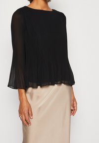 Schwarze plissierte Bluse mit langen Glockenärmeln und einem Boot-Ausschnitt, kombiniert mit einem beigen Satinrock. Weiche, fließende Stofftextur.