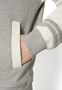 Handen in framfickan på en grå och vit långärmad sweatshirt med ribbad mudd och randig ärmdetalj.