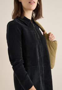 Giacca di cotone a coste nera con chiusura a zip, texture prominente e maniche lunghe; il modello tiene una borsa piegata di colore verde oliva.