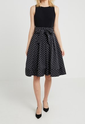 Vrouw die een mouwloze zwarte jurk draagt met een uitlopende rok met polkadots en een strik in de taille, gecombineerd met zwarte puntige hakken.