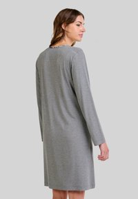 Mujer vistiendo un vestido gris de manga larga y hasta la rodilla, de pie con la espalda parcialmente girada contra un fondo gris liso.