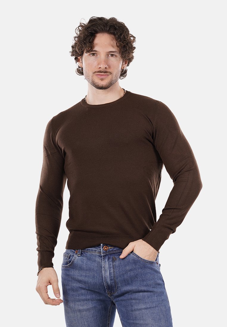 Uomo con capelli ricci che indossa una camicia a maniche lunghe marrone e jeans blu, in piedi con una mano in tasca su uno sfondo semplice.