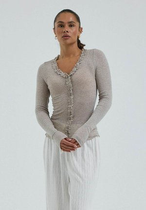 Donna con capelli scuri raccolti in una coda di cavallo, indossa una maglia grigia aderente a maniche lunghe con volant e pantaloni bianchi con fantasie, in piedi davanti a uno sfondo semplice.