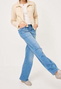 Jasnobeżowa kurtka na białej bluzce, połączona z jasnoniebieskimi dzwonami i szarymi trampkami. Kurtka ma guziki i szczegół ze sznurkiem.