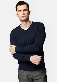 Granatowy sweter w serek wykonany z miękkiego materiału, z dopasowanymi długimi rękawami oraz ribbami przy mankietach i dolnym wykończeniu. Subtelny detal logo w okolicy klatki piersiowej.