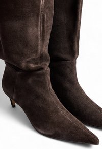 Stivali alti fino al ginocchio in camoscio marrone con punta affusolata e tacco basso. Presentano un design morbido nella parte superiore con dettagli di cucitura visibili.