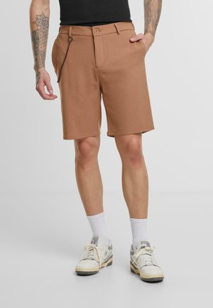Brown tailored shorts with a flat front, side pockets, and a metal chain accent. Paired with white socks and sport shoes.