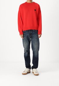 Pull en maille rouge à encolure ronde avec un petit logo cœur noir, associé à un jean bleu marine et des baskets blanches.