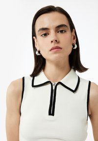 Vit ärmlös topp med svarta detaljer och en dragkedjekrage. Ribbstruktur med en figursydd design. Silverfärgade hoopörhängen.