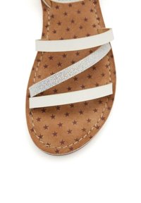 Sandalia blanca de tiras con una correa de purpurina plateada, con una suela marrón que presenta un patrón de estrellas. Cuenta con un borde cosido y una textura suave.