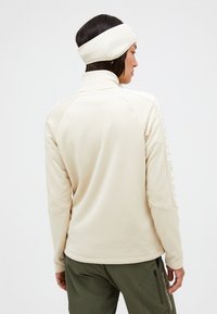 Beige Fleecejacke mit hohem Kragen, tailliertem Design und sichtbarem Branding am linken Ärmel. Kombiniert mit grünen Hosen.