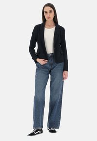 Blazer nero su una camicia bianca, abbinato a jeans blu a vita alta e a gambale largo e sneakers nere. Tessuto liscio, design strutturato.