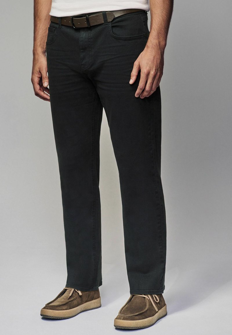 Jeans de mezclilla negra con corte recto, complementados con un cinturón marrón. Combinados con zapatos de ante marrón con un acabado texturizado.
