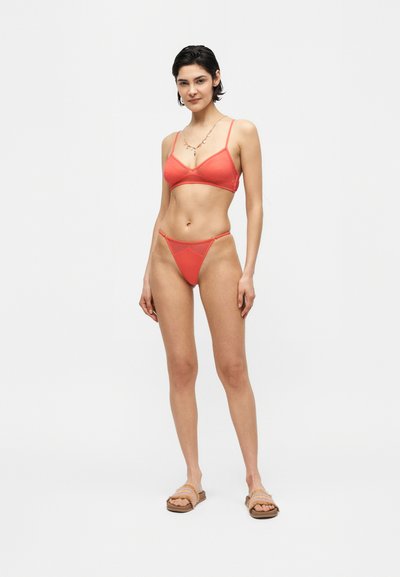 Γυναίκα που φοράει κοράλλι μπικίνι και σανδάλια σε απλό φωτεινό φόντο, φορώντας κολιέ.