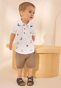 Weißes Poloshirt mit mehrfarbiger, nautischer Stickerei, braune Shorts und blaue Sandalen mit Schnallen, neben einem Holztisch stehend.