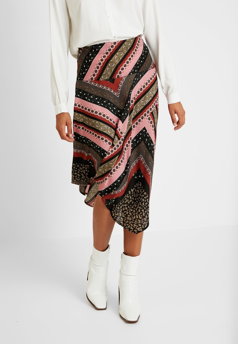 Femme portant une jupe midi asymétrique à motifs multiples avec des rayures roses, noires, marron et rouges, assortie à des bottines blanches à talons.