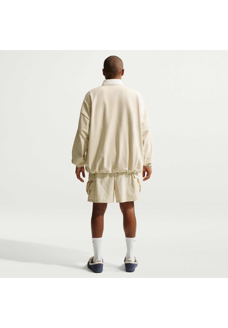 Homme debout, dos à la caméra, portant un sweat crème ample, un short assorti, des chaussettes blanches et des sandales sur un fond uni.
