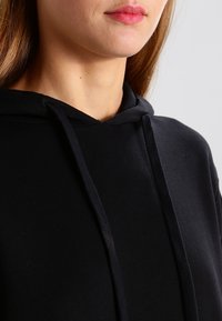 Svart huvtröja med dragsko i halsen, med en slät textur och en avslappnad passform. Närbild visar tyg- och sömnadsdetaljer.