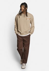 Beige sweatshirt med et grafisk design på brystet, parret med brune bukser og hvide sneakers. Personen bærer en sort strikbeanie.