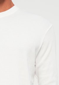 Close-up van een persoon die een eenvoudig wit geribbeld longsleeve shirt met een ronde hals draagt, tegen een neutrale achtergrond.
