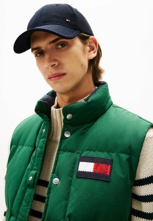 Padded green vest with a high collar, featuring a patch of red, white, and navy blue. Worn over a cream cable-knit sweater and a navy cap.