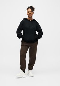 Jeune femme debout, les mains dans les poches de son sweat à capuche noir, portant un pantalon de jogging marron et des baskets blanches sur un fond blanc uni.