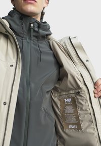 Bež vanjska jakna preko tamnozelene unutarnje jakne. Značajke uključuju visoki ovratnik, patentne zatvarače i oznaku koja prikazuje upute za njegu.