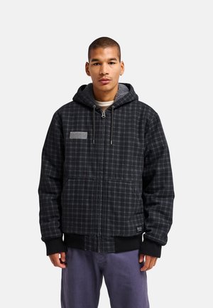 Jeune homme portant une veste à capuche à carreaux noirs et gris avec poches avant et fermeture éclair, debout devant un fond clair uni.
