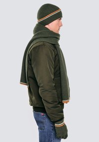 Oliwkowa dzianinowa czapka z pomarańczowymi i białymi paskami, dopasowany szalik i rękawiczki. Noszone z ciemną kurtką i niebieskimi dżinsami. Widok z boku.