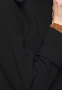 Schwarzer Stoff mit einem leichten Glanz, der eine weiche Textur aufweist und einen gerollten Ärmel mit braunem geripptem Akzent am Bund hat. Schlichte Designelemente.